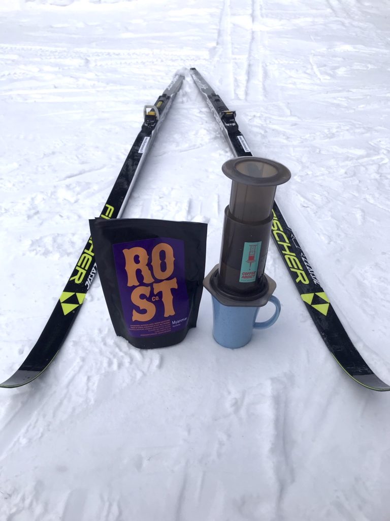 Sukset, kahvipussi, aeropress ja muki lumessa.
