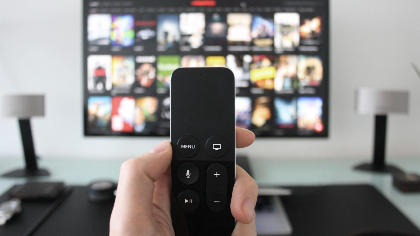 Lähikuvassa käsi osoittaa Apple TV:n Siri Remote -kaukosäätimellä televisiota, jossa on auki suoratoistopalvelun elokuvien ja sarjojen kansikuvagalleria.
