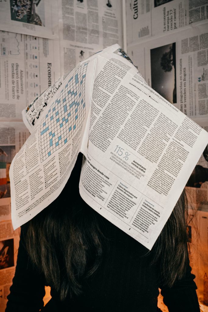 Pitkähiuksinen ihminen sanomalehti päänsä päällä peittäen kasvot ja päälaen.