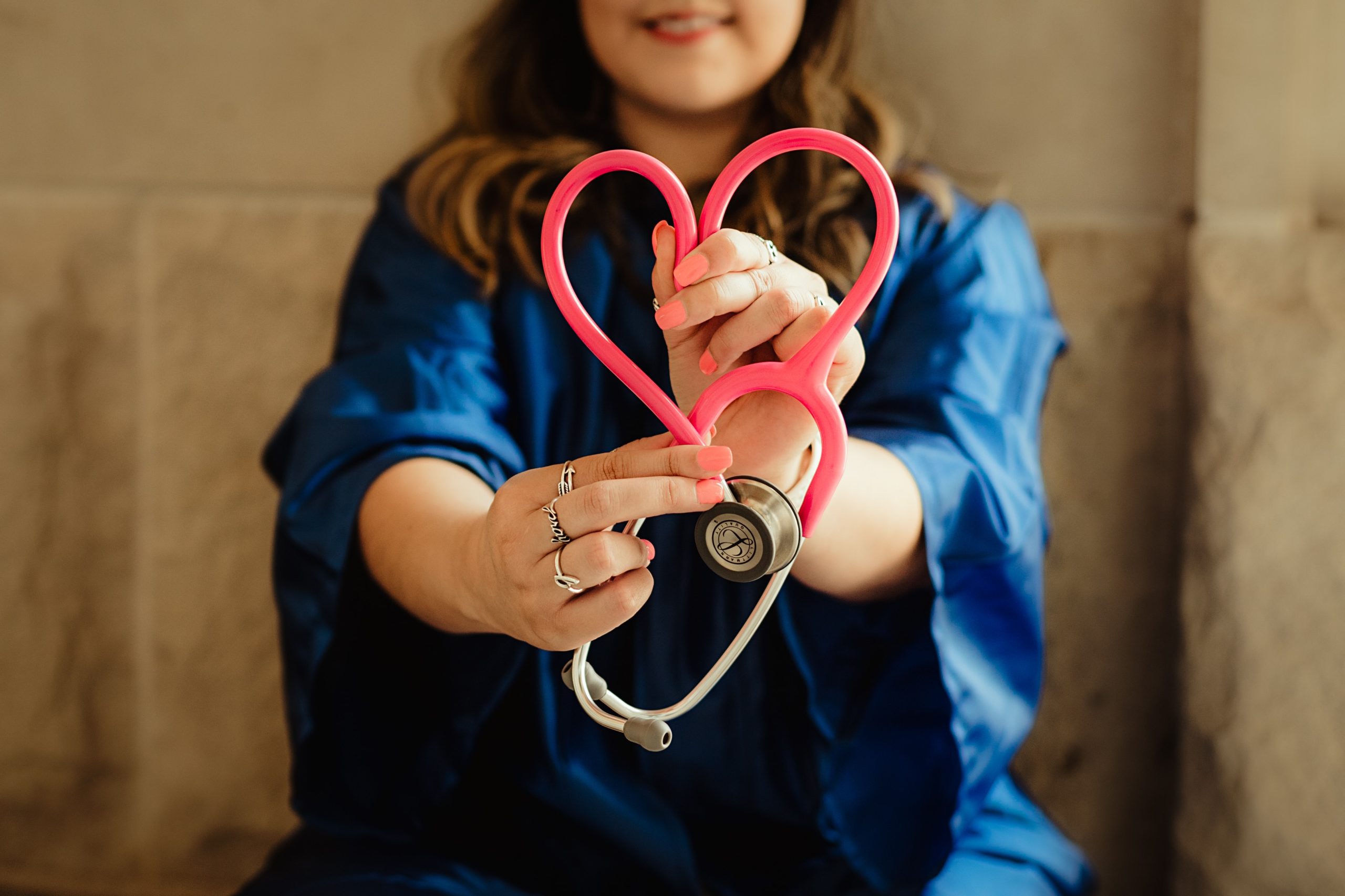 Kuvassa näkyy työvaatteisiin pukeutunut hoitaja tai lääkäri, joka pitelee käsissään stetoskooppia. Stetoskoopin pinkki johto on taitettu sydämen muotoiseksi kuvioksi.